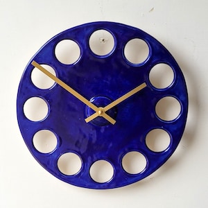 Op de afbeelding: Een blauwe keramische klok met gouden wijzers en een uniek ontwerp met twaalf ronde uitsparingen rond de wijzerplaat.