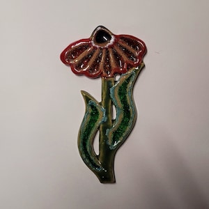Può includere: Un appendiabiti da parete in ceramica a forma di fiore con un fiore rosso e marrone e foglie verdi. Il fiore ha un centro nero.