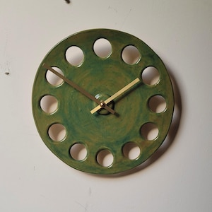 May include: A round, green ceramic clock with a series of circular cutouts around the perimeter. The clock face has a glossy finish and gold-colored hands. The clock is mounted on a white wall.