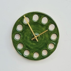 Op de afbeelding: Een ronde wandklok met een glanzend donkergroen geglazuurde keramische rand, voorzien van een reeks gelijkmatig verdeelde ronde uitsparingen langs de omtrek. De wijzerplaat heeft dunne, goudkleurige metalen uur-, minuut- en secondewijzers.