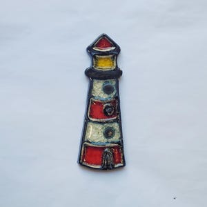 Op de afbeelding: Een keramisch vuurtorenornament met een glanzende afwerking en een zwarte omlijning. De structuur toont afwisselende horizontale banden van rood en wit craquelé glazuur, een gele lantaarn, een rood dak en kleine zwarte ronde vensters.