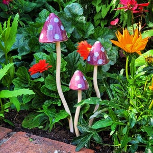 May include: Three ceramic mushroom garden decorations with white polka dots. The mushrooms are pink and white and have a rustic, whimsical style. They are displayed in a garden setting with green foliage and flowers.