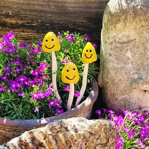 May include: Three yellow ceramic mushrooms with smiley faces are arranged in a terracotta pot filled with purple flowers. The mushrooms are painted white and have a stem.