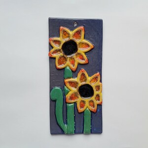 Peut inclure: Une suspension murale en céramique avec deux tournesols sur fond bleu. Les tournesols sont jaunes avec des accents orange et rouge. Les tiges sont vertes.