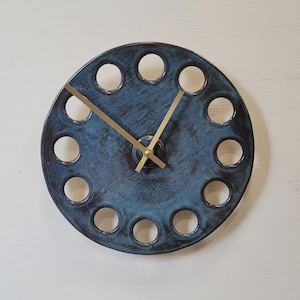 May include: A circular ceramic wall clock with a mottled dark blue and black glaze. The clock face features twelve evenly spaced circular cutouts along the perimeter and three thin, gold-coloured metal hands centred on the dial.