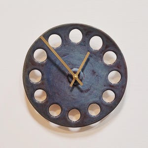 Può includere: Un orologio rotondo in ceramica blu con una serie di fori distanziati. L'orologio ha lancette dorate e un centro. Un oggetto decorativo per la parete.