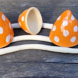Może przedstawiać: Trzy pomarańczowe ceramiczne grzyby w białe kropki. Jeden grzyb jest otwarty, ukazując wnętrze. Grzyby są ułożone na białej, falistej, ceramicznej podstawie.