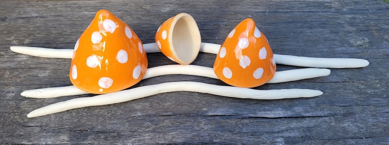 Może przedstawiać: Trzy pomarańczowe ceramiczne grzyby w białe kropki. Jeden grzyb jest otwarty, ukazując wnętrze. Grzyby są ułożone na białej, falistej, ceramicznej podstawie.