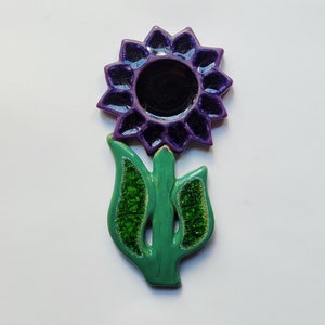 Może przedstawiać: Ceramiczny kwiat w kolorze fioletowym z zielonymi liśćmi. Kwiat ma ciemnofioletowy środek i jest wykonany z glazurowanej ceramiki.