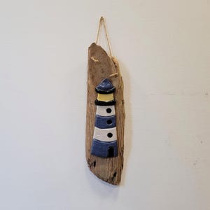May include: A wall hanging featuring a ceramic lighthouse with blue and white horizontal stripes, a yellow light section, and black accents, mounted on a piece of natural driftwood with a thin jute twine loop for hanging on a wall.
