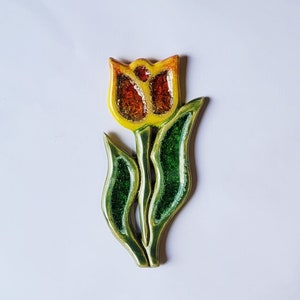 Puede incluir: Una tulipa de cerámica amarilla y naranja con hojas verdes. La tulipa tiene una superficie texturizada y es una pieza decorativa.