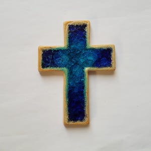 May include: A handmade cross with a light brown border and a vibrant blue center. The blue center has a textured, crystalline appearance, with shades ranging from light turquoise to deep indigo. The cross is a religious symbol.