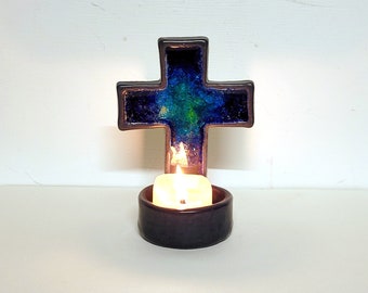 Könnte beinhalten: Ein Kerzenhalter aus Keramik in Form eines Kreuzes mit einer teal- und blauen Glasur. Der Halter hat eine kleine Kerze im Inneren, die brennt.