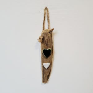 Könnte beinhalten: Ein rustikaler Wandbehang aus Treibholz mit einem natürlichen Seilaufhänger. Das Holzstück hat einen herzförmigen Keramikeinsatz in Schwarz und einen herzförmigen Keramikeinsatz in Weiß. Das Treibholz hat eine natürliche braune Farbe.