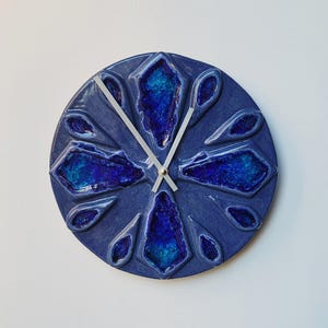 May include: A round, blue ceramic wall clock with a decorative design. The clock face features a dark blue background with raised, geometric shapes in varying shades of blue and silver clock hands.