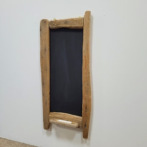 May include: A rectangular chalkboard with a rustic, weathered wooden frame. The chalkboard is black, and a piece of white chalk rests in a small wooden holder at the bottom. The frame is made of light brown, rough-hewn wood.