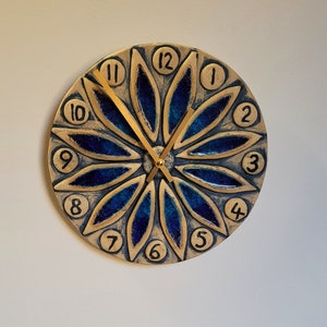 May include: A blue and gold ceramic clock with a flower design and gold hands. The numbers 1 through 12 are displayed on the clock face.
