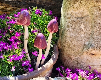 May include: Three purple ceramic mushrooms with white stems are planted in a terracotta pot filled with purple flowers. The pot is sitting on a stone surface next to a large, textured stone.