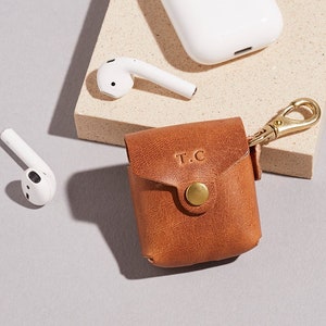 May include: A brown leather case with a gold clasp and keychain. The case has the initials "T.C." embossed on the front. The case is designed to hold a pair of wireless earbuds.
