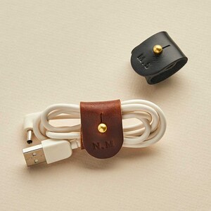 May include: Two leather cord organisers with gold accents. One is brown with the initials "N.M." and the other is black with the initials "M.S."