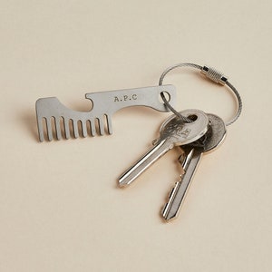 May include: A silver key chain with a comb and bottle opener attached. The keychain has the text "A.P.C." engraved on it. Two silver keys are attached to the keychain.