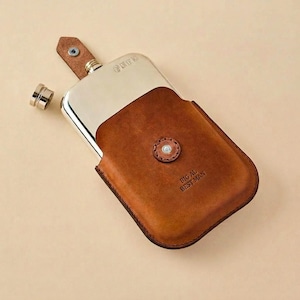 May include: A rectangular metal flask encased in a brown leather sleeve with a snap button closure. The leather is embossed with the text BIG AL BEST MAN. A matching leather tab with a metal rivet extends from the top, and a small metal cap sits nearby.