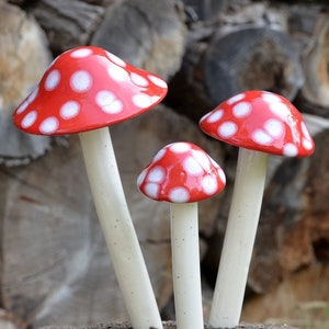 May include: Three ceramic mushroom sculptures with glossy red caps featuring white polka dots and smooth, off-white cylindrical stems. The mushrooms are arranged in a cluster, with one large, one medium, and one small mushroom standing upright.