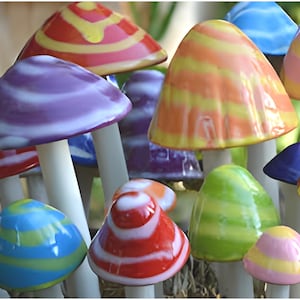 May include: A collection of ceramic mushroom sculptures featuring glossy, multi-colored caps with various striped and swirled patterns in shades of red, orange, yellow, green, blue, and purple, each supported by a smooth, cylindrical white stem.