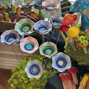 May include: A collection of colorful ceramic flower-shaped bowls and mushroom sculptures. The bowls have various colors, including pink, blue, and green, with intricate spiral and floral designs in the center. The mushrooms are painted in bright colors.