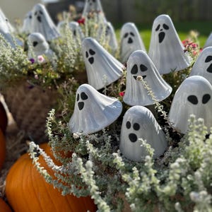 May include: A collection of white ceramic ghost figurines with black eyes and mouths, nestled among green plants and orange pumpkins. The ghosts have a draped sheet design, perfect for Halloween decorations.