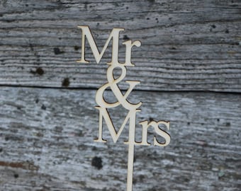 Puede incluir: Un adorno de pastel de madera con las palabras "Mr & Mrs" en una fuente clásica. El adorno está colocado sobre un pastel glaseado decorado con chispas doradas y pequeñas flores. El fondo es una superficie de madera envejecida.