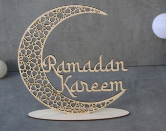 May include: A wooden crescent moon cutout with an intricate geometric pattern and the words "Ramadan Kareem" inscribed on it. The moon is standing on a wooden base.