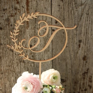 Peut inclure: Un topper de gâteau en bois avec une lettre cursive "T" dans un cercle entouré de feuilles. Le topper est placé sur un gâteau de mariage blanc décoré de fleurs roses et blanches.