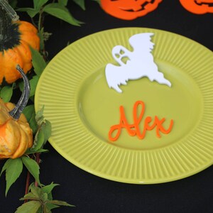 Può includere: Un piatto verde con un fantasma bianco e il nome "Alex" scritto in arancione. Il piatto è su una superficie nera con una vite verde e piccole zucche arancioni.