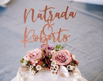 Puede incluir: Un pastel redondo, blanco y glaseado, sobre un soporte blanco para pasteles, decorado con rosas rosas y vegetación. Un adorno de pastel de madera dice "Natsada & Roberto". El pastel es una pieza central para una celebración.