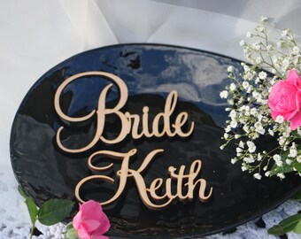 May include: Two wooden signs with the words "Bride" and "Keith" in cursive script on a black background. The signs are on a black plate with white flowers in the background.