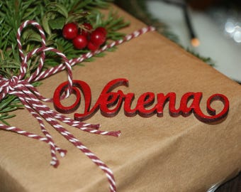Puede incluir: Un regalo envuelto en papel marrón, atado con hilo rojo y blanco. El regalo está decorado con ramitas de hoja perenne, bayas rojas y una etiqueta de madera roja con el nombre "Verena". Un tema festivo de vacaciones.