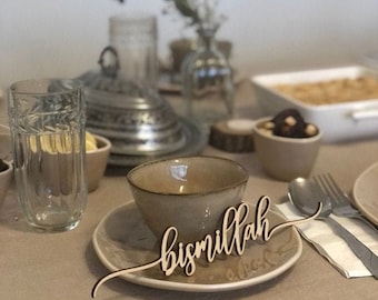 May include: A wooden sign with the word "bismillah" written in cursive script. The sign is placed on a table setting with a bowl, a glass of water, and a napkin.