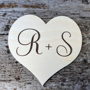 May include: A wooden heart-shaped decoration with the engraved letters "R" and "S" separated by a plus sign. The heart is light-coloured, and the letters are a darker brown. The background is a weathered wooden surface.