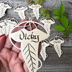 Peut inclure: Des étiquettes de nom en forme de feuille en bois clair, gravées de noms tels que "Vicky" et "David" dans une police stylisée. Les étiquettes présentent un motif de nervures de feuilles. Des feuilles et des vignes vertes sont en arrière-plan.