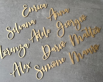 May include: Gold-colored wooden name cutouts in various cursive fonts. Names include Enrica, Anna, Silvana, Giorgio, Adele, Matteo, Lorenza, Dario, Alex, Simone, and Mauro. Ideal for wedding place settings or personalized gifts.