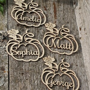 Puede incluir: Cuatro porta tarjetas de lugar de madera en forma de calabaza con los nombres "Amelie", "Sophia", "Matt" y "George" escritos en ellos.