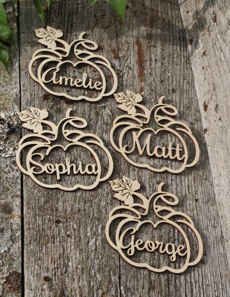 Puede incluir: Cuatro porta tarjetas de lugar de madera en forma de calabaza con los nombres "Amelie", "Sophia", "Matt" y "George" escritos en ellos.