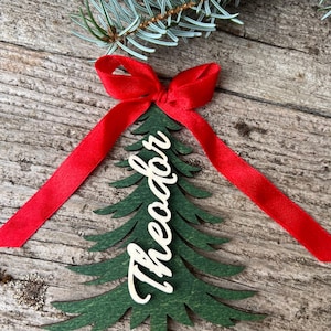 Peut inclure: Ornement de Noël en bois vert en forme de sapin, avec le nom "Theodor" en écriture cursive. Il est orné d'un nœud et d'un ruban en satin rouge. Le fond est une surface en bois rustique avec une branche de conifère.