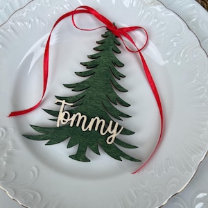 Puede incluir: Tarjeta de lugar con forma de árbol de Navidad de madera verde con el nombre "Tommy" en cursiva, atada con una cinta roja, sobre un plato blanco con borde dorado. El plato tiene un patrón en relieve.