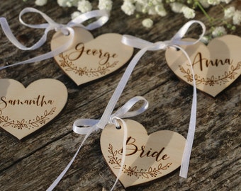 May include: Four wooden heart-shaped tags with white ribbon, each with a different name engraved in cursive: Samantha, George, Anna, and Bride. The tags are on a wooden surface.