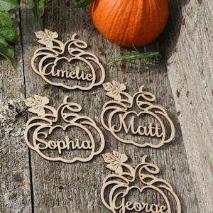 Puede incluir: Cuatro porta tarjetas de madera en forma de calabaza con los nombres Amelia, Sophia, Matt y George. Las tarjetas est&aacute;n sobre una superficie de madera con una peque&ntilde;a calabaza naranja.