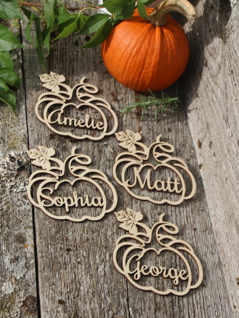 Puede incluir: Cuatro porta tarjetas de madera en forma de calabaza con los nombres Amelia, Sophia, Matt y George. Las tarjetas est&aacute;n sobre una superficie de madera con una peque&ntilde;a calabaza naranja.