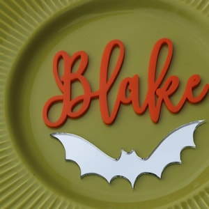 Può includere: Un piatto verde con un pipistrello bianco e il nome "Blake" scritto in arancione.