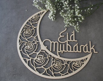 May include: A wooden crescent moon decoration featuring a rose design and the Arabic phrase "Eid Mubarak". The light-coloured wood moon is set against a grey backdrop, with white flowers in the top right corner. A decorative item for celebrations.